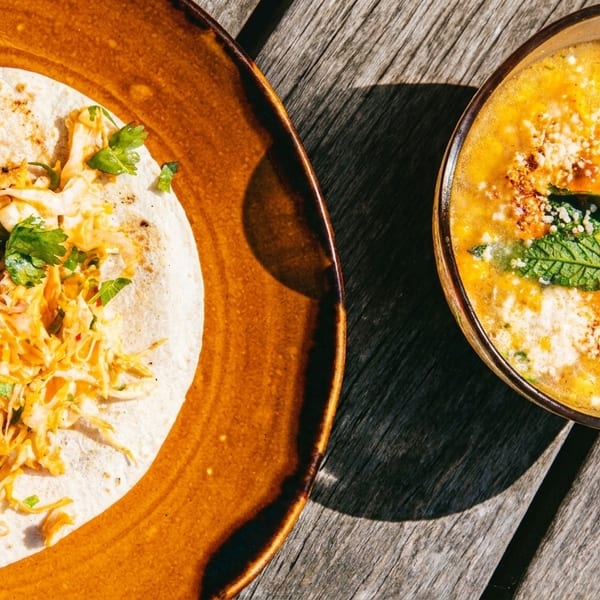 Shrimp taco with slaw on a brown plate next to a bowl of corn soup with mint.