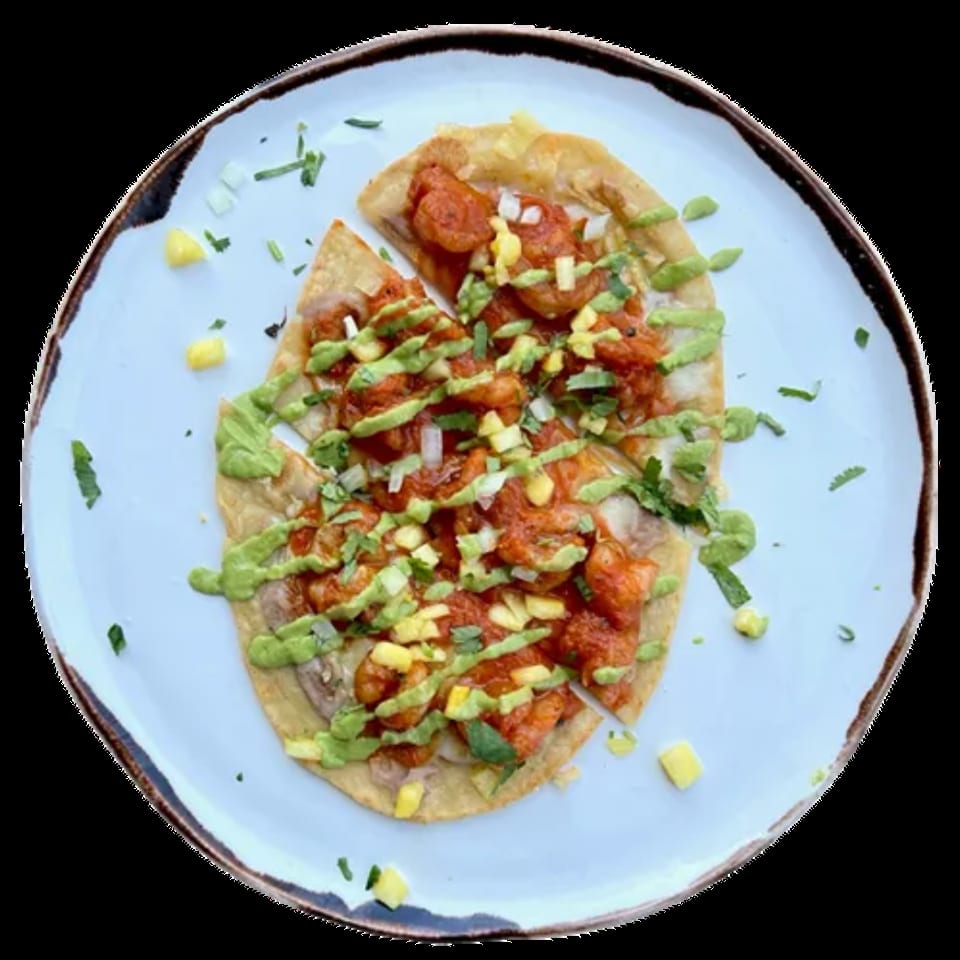 Overhead view of a shrimp tostada with red sauce, green drizzle, and diced mango on a white plate.